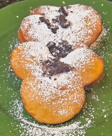 Beignets From Beyond Recipe: 3 Mickey shaped beignets on a green on top of cookies and cream cheese icing with powdered sugar and crushed oreos on top.